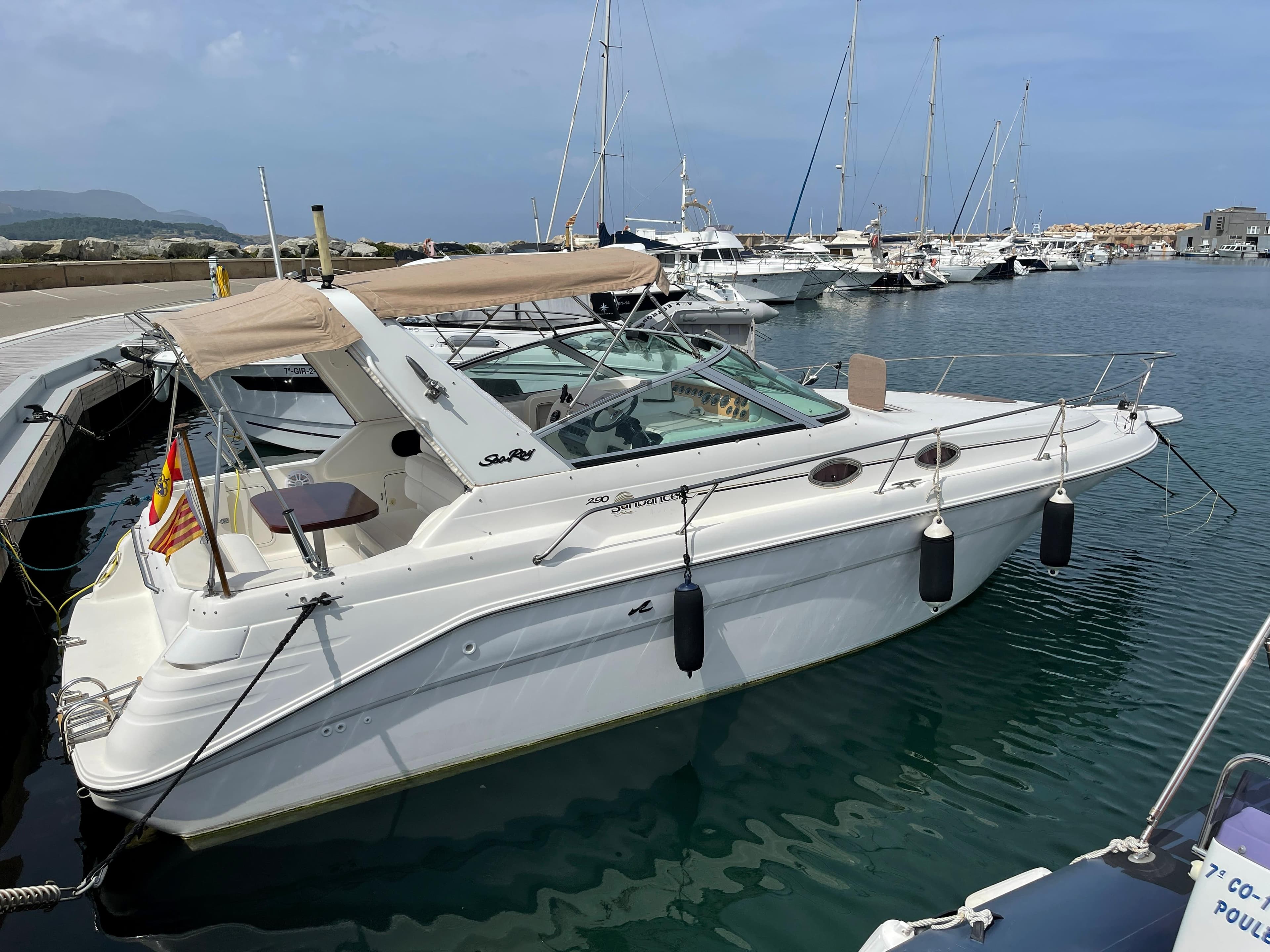 A 1997 Sea Ray 290 Sundancer docked in a marina. It features a beige canopy and fenders.